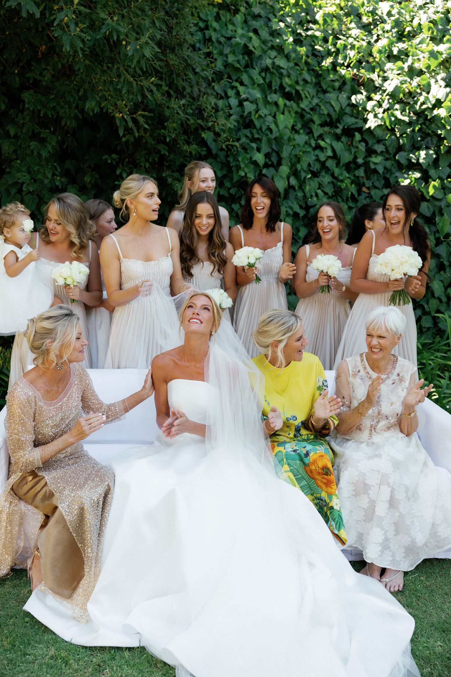 Wedding party in light neutral dresses laughs together during outdoor photo session in garden setting.