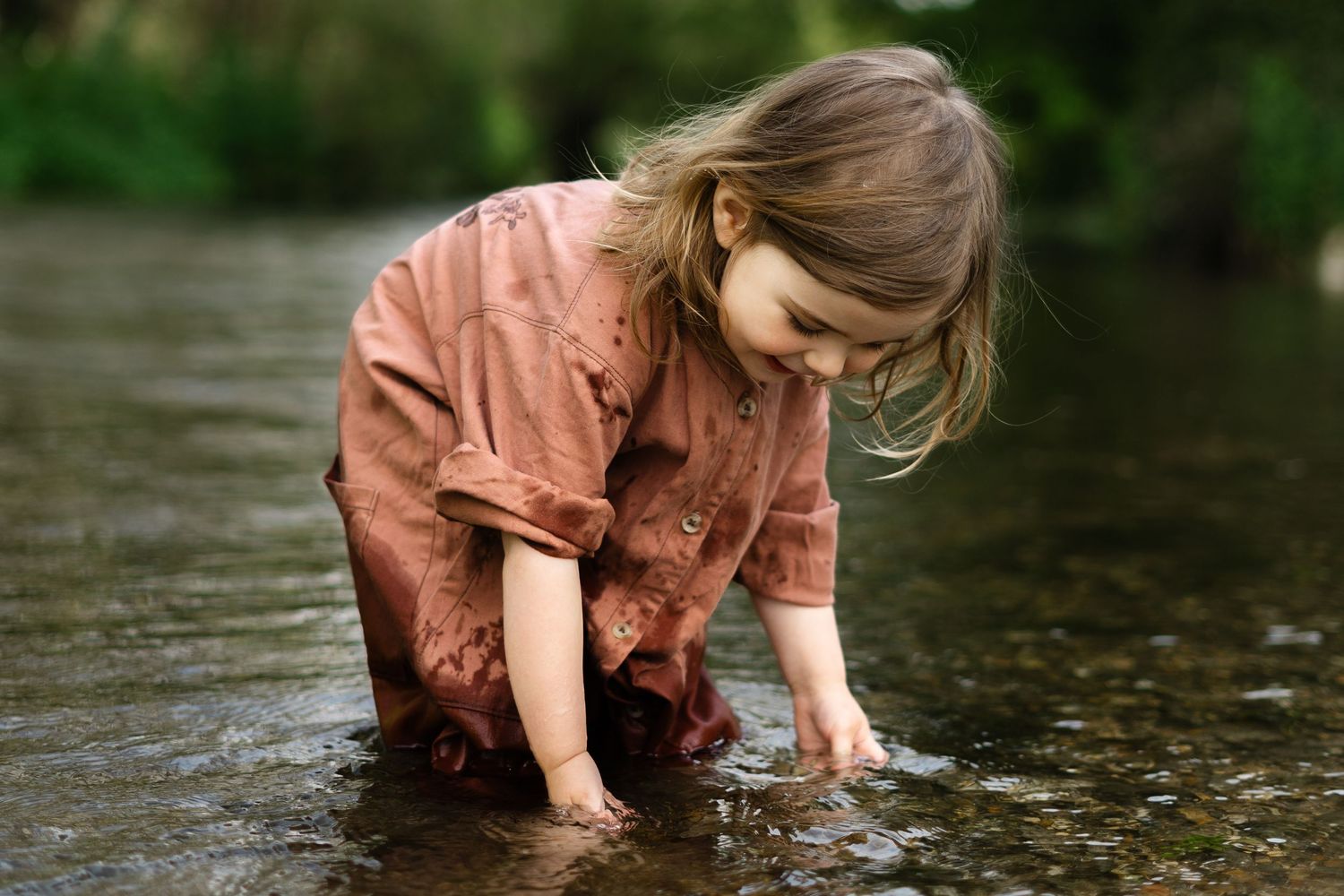 Child in a pink dress plays in a shallow stream, exploring the flowing water on a summer day.
