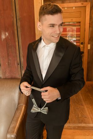 Person in black tuxedo adjusting their tie while standing in a wooden interior.