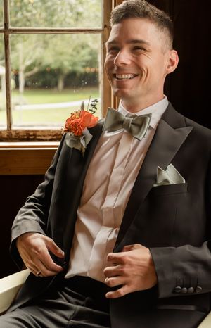 Groom in formal attire with coral boutonniere smiling by window in rustic setting.