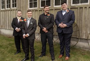Four groomsmen in formal attire stand together outside a rustic building on a wedding day.