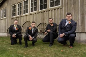 Row of groomsmen crouching in casual poses outside a rustic wooden building.