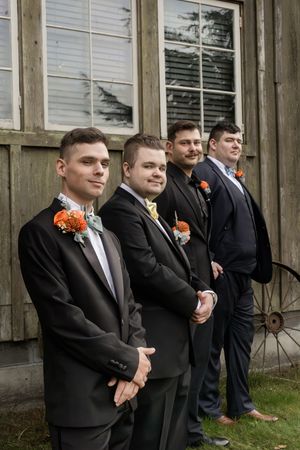 Groomsmen in black suits with orange boutonnieres stand in line against a rustic wooden wall.