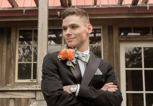 Groomsmen in dark suits pose casually against a rustic barn backdrop with orange floral accents.