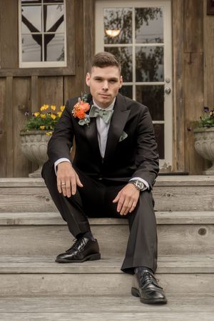 Groom in classic tuxedo with coral boutonniere poses on wooden steps of rustic building.