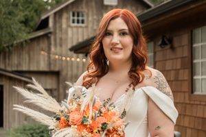 A bride in a white dress holds a vibrant orange and coral bouquet with pampas grass against a rustic barn backdrop.