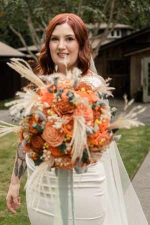 A bridal bouquet features orange roses, coral flowers and dried pampas grass elements in a modern arrangement.