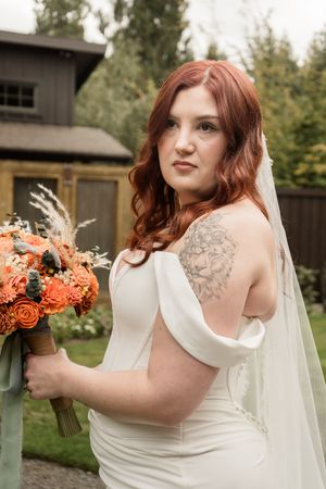 A bride in a sleeveless white gown poses with an autumn-themed bouquet featuring orange blooms and dried elements.