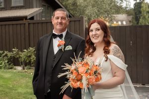 Happy couple poses outdoors with coral bouquet against rustic wooden building backdrop.