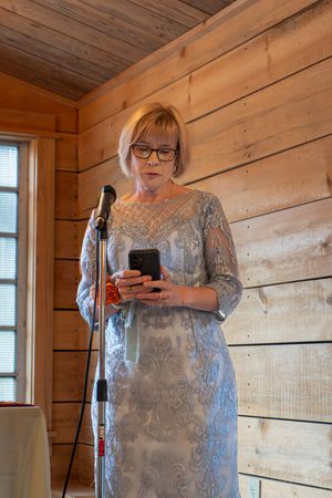 Speaker in lace dress holds phone at wooden podium during wedding reception.