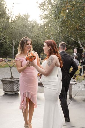 Two women in pink and white dresses share a joyful moment at an outdoor wedding celebration.
