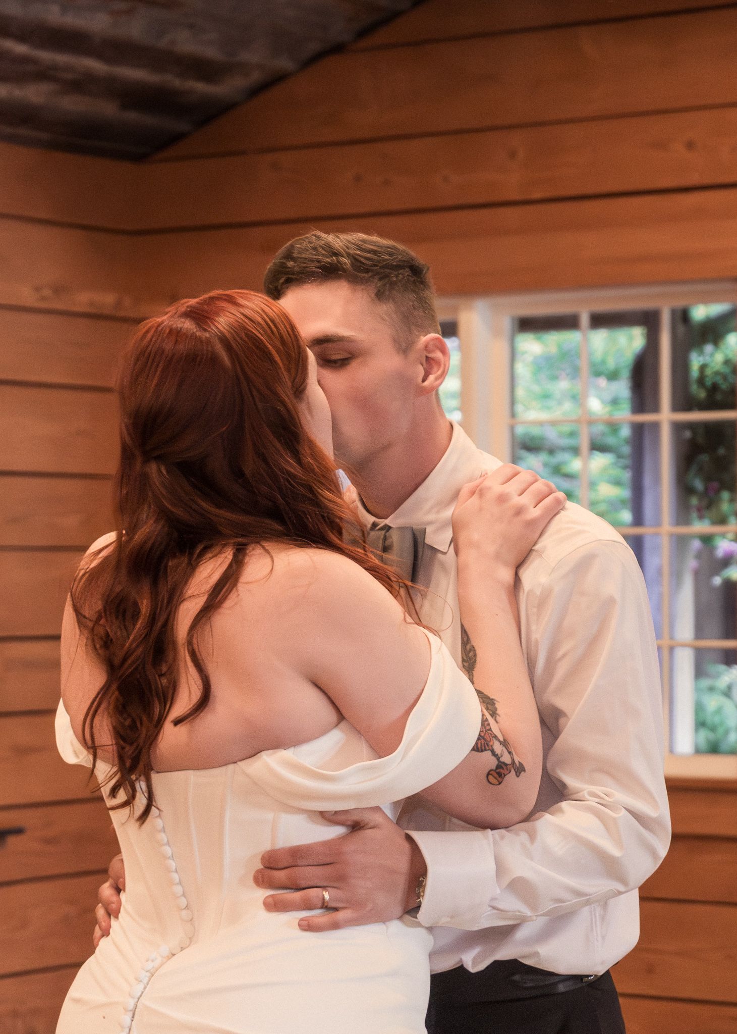 A romantic wedding kiss between newlyweds in front of a rustic wooden cabin wall.