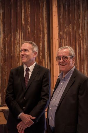Group of men in formal attire gathered in front of wooden wall panels during an event.