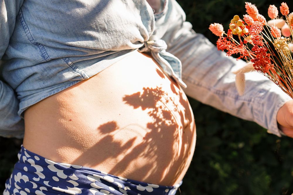Shooting de grossesse en extérieur par photographe de famille à Beersel. Lumière naturelle. Ventre nu avec reflets du bouquet de fleurs.