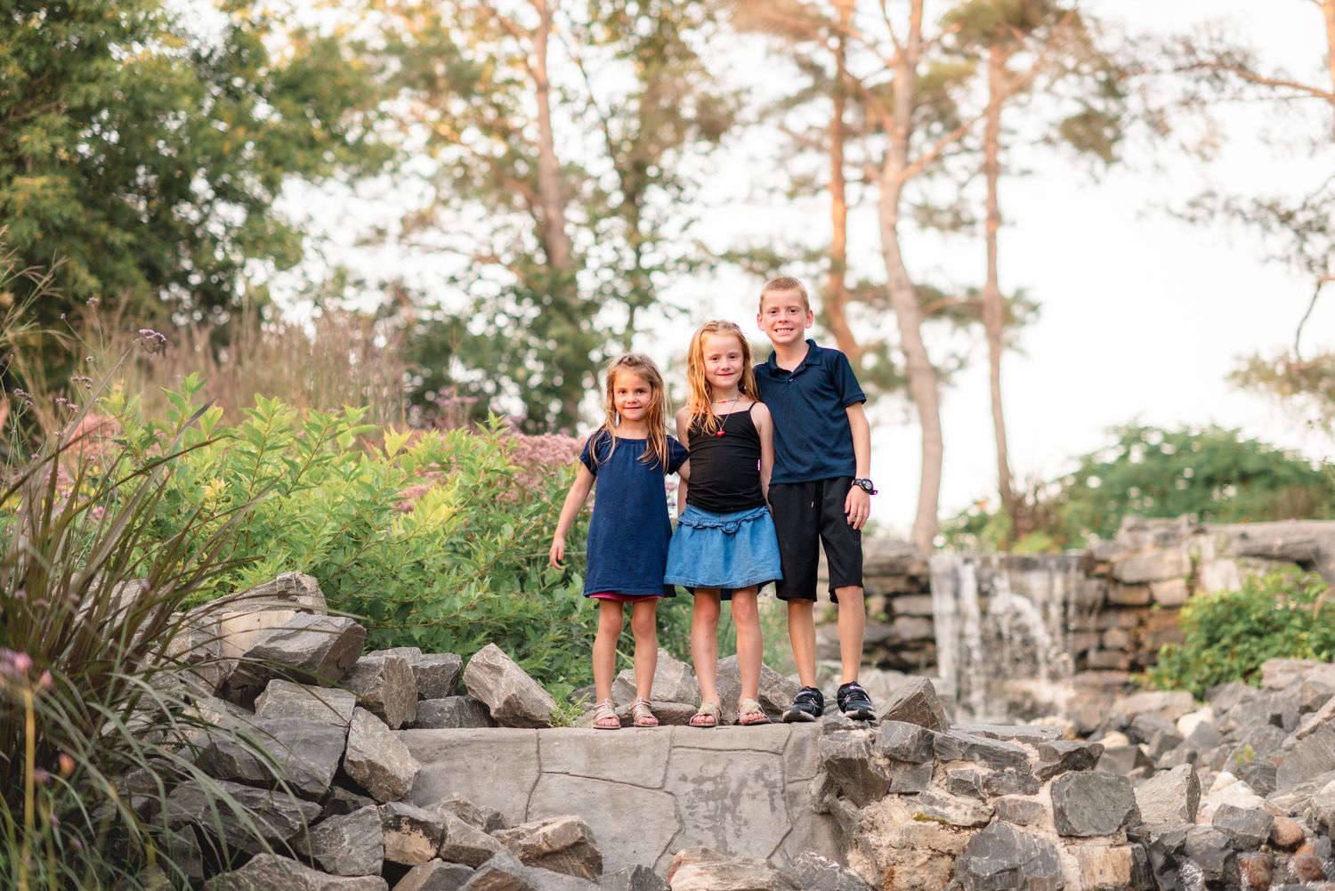 Children in matching blue outfits sit on a rustic wooden stump during golden hour in a lush garden setting.