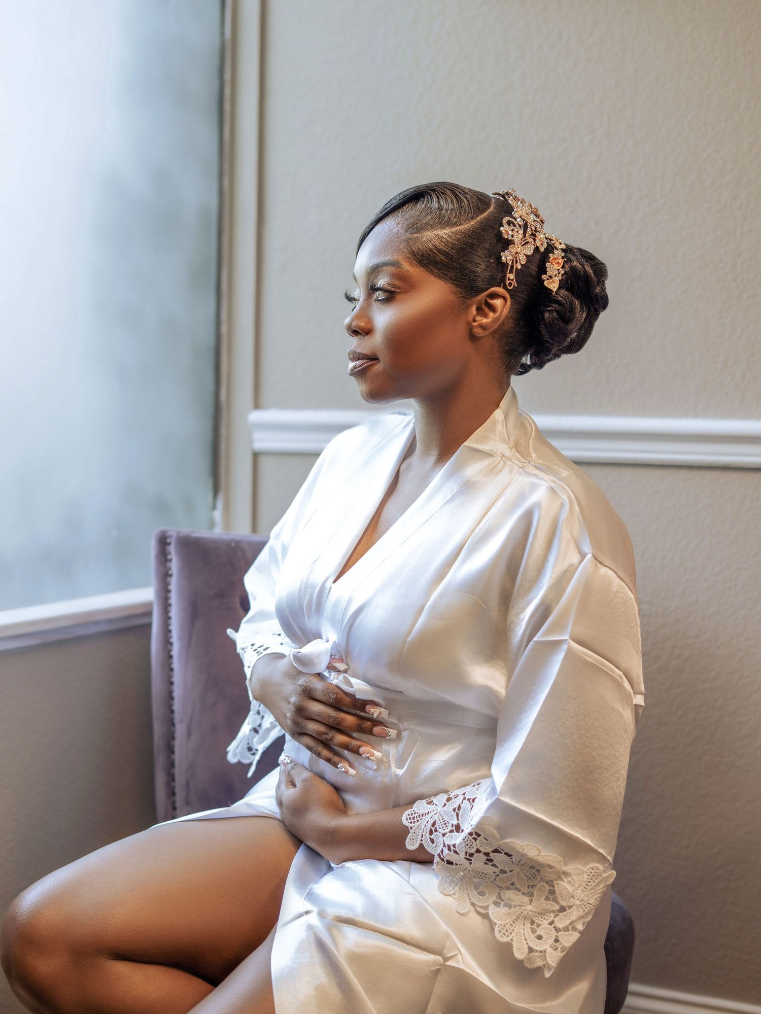 A bride in a white satin robe with lace trim sits in profile against a neutral wall.
