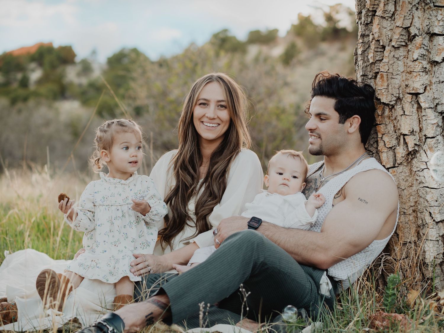 Family relaxing together outdoors on grass near a tree trunk.