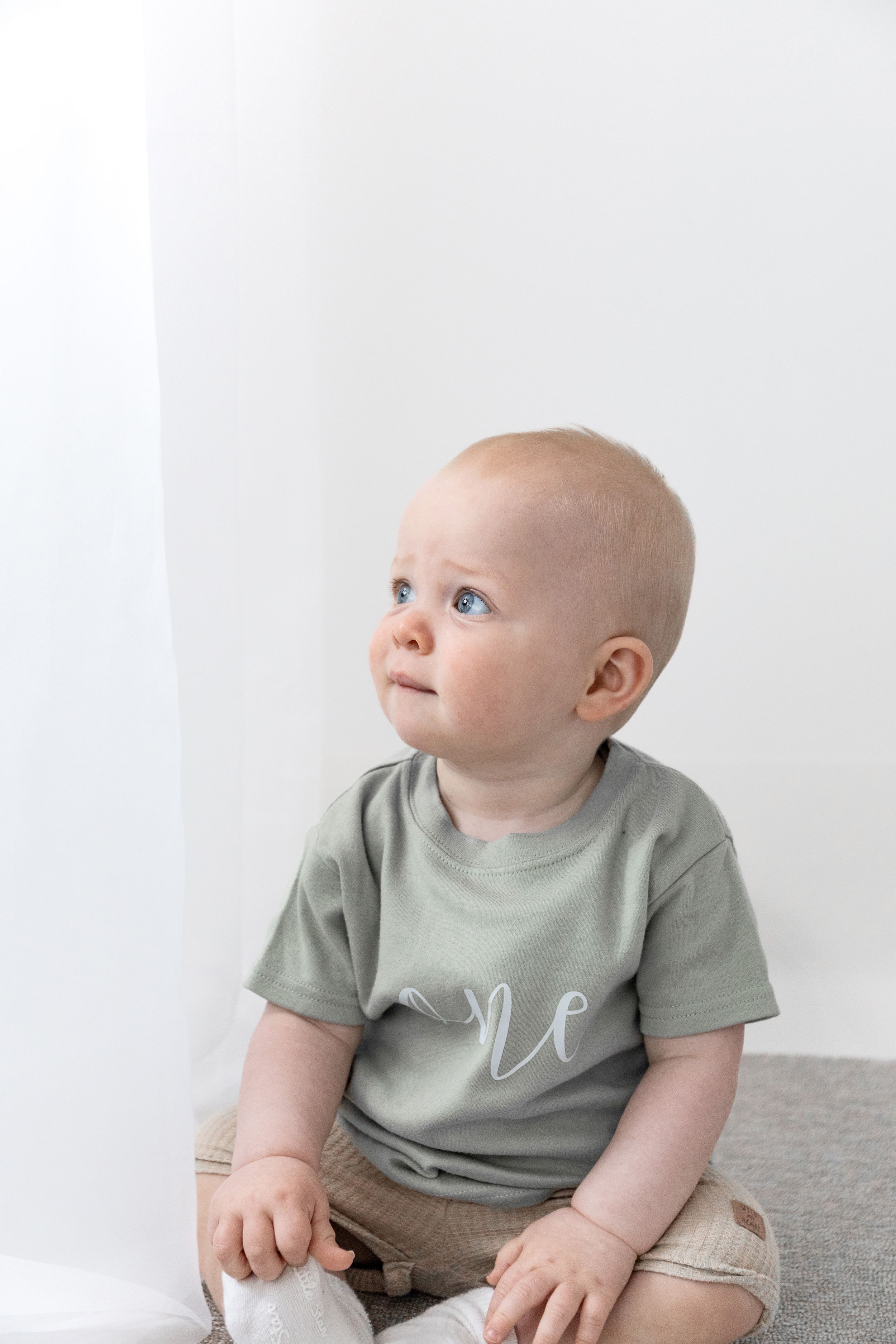 A baby in a sage green "one" t-shirt sits on a light surface looking upward with curiosity during birthday shoot in Nottingham