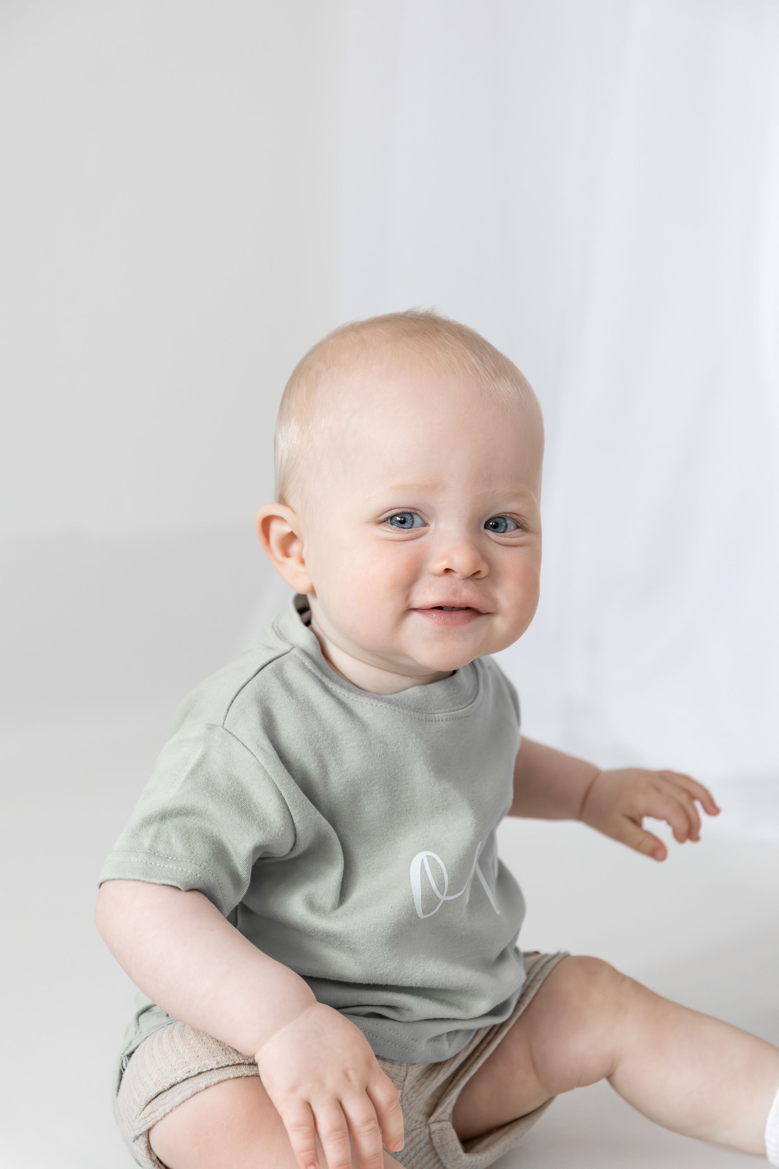 Baby in mint green t-shirt sits and smiles against white background during birthday shoot in Nottingham.