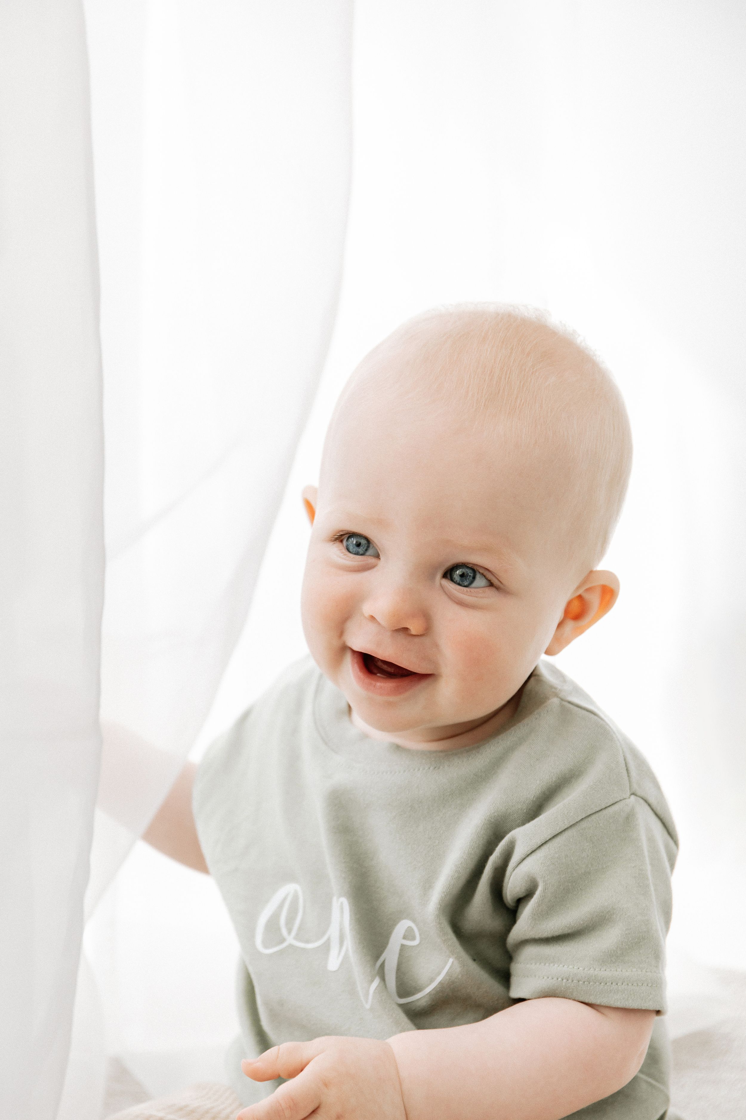 Happy baby wearing a light green t-shirt sits by white curtains in a bright room, during birthday shoot in Nottingham