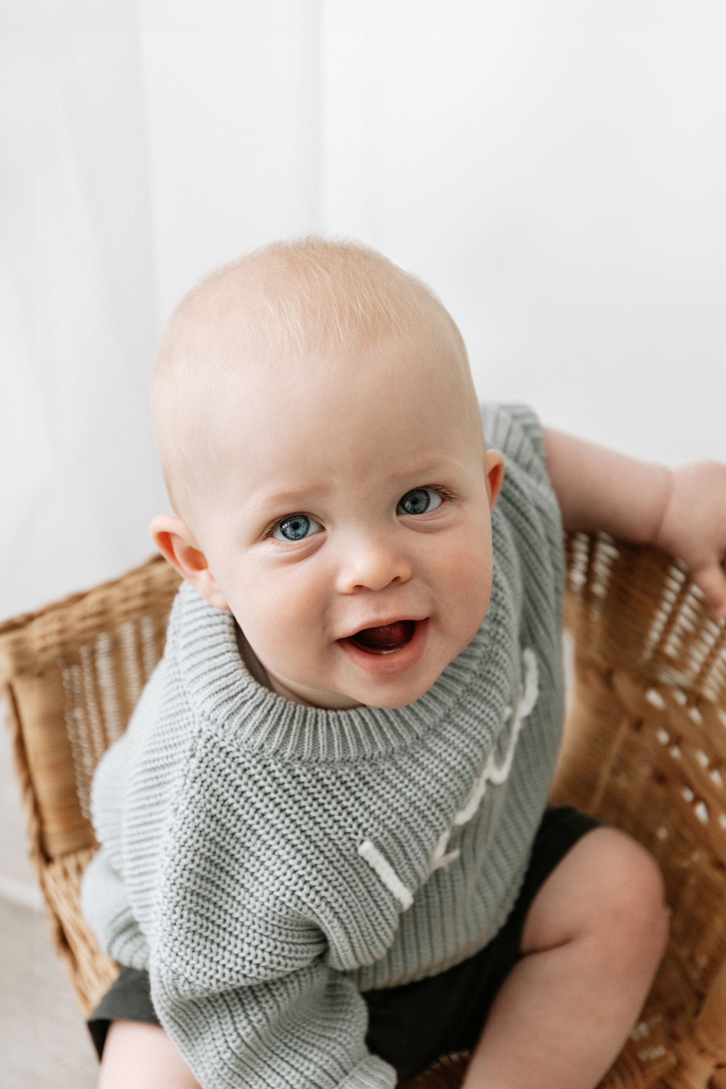 A happy baby wearing a blue knit jumper sits in a wicker chair against a white background, during birthday shoot in Nottingham
