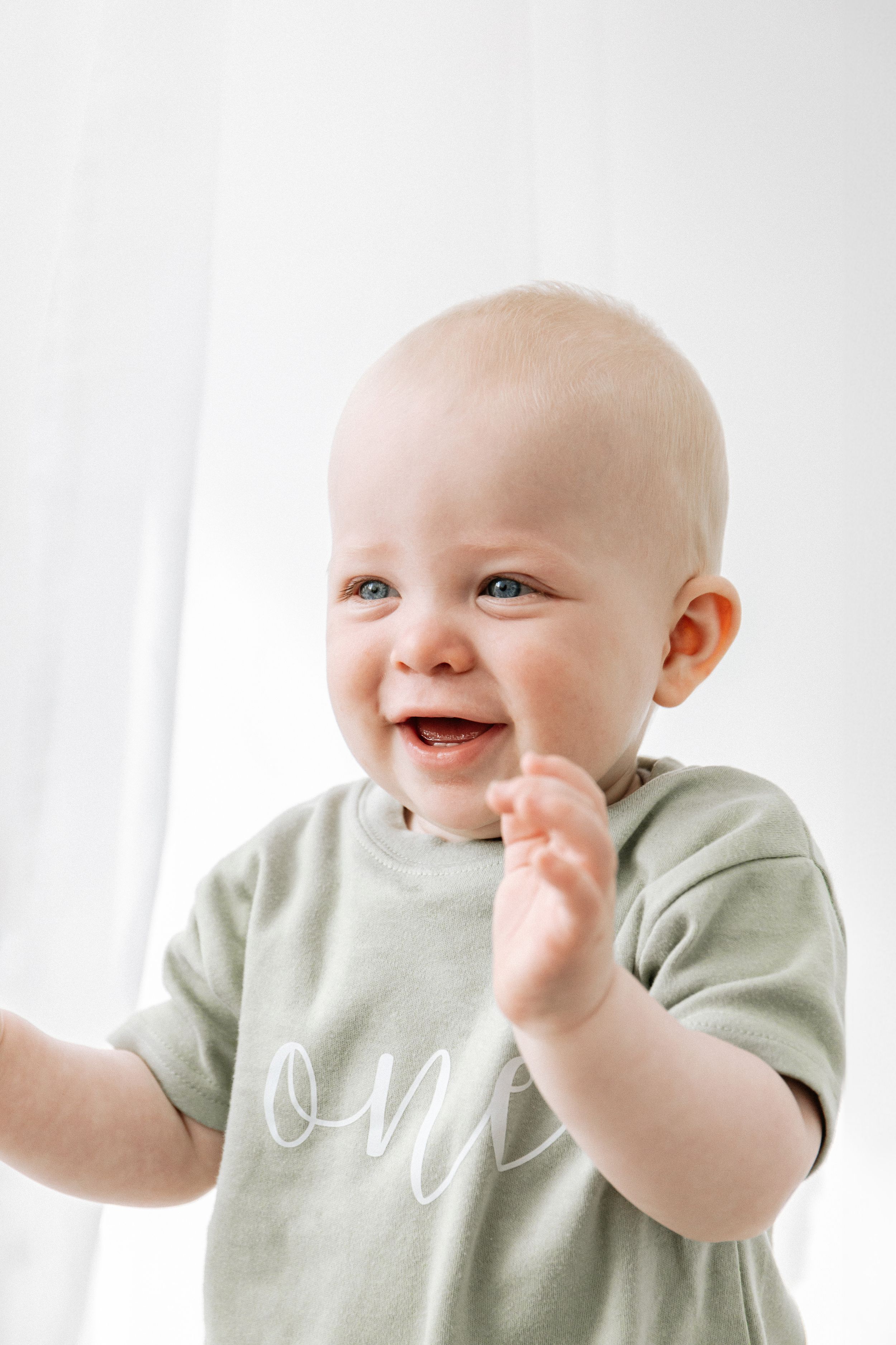 Baby in light green t-shirt laughs and waves joyfully against white background, during birthday shoot in Nottingham