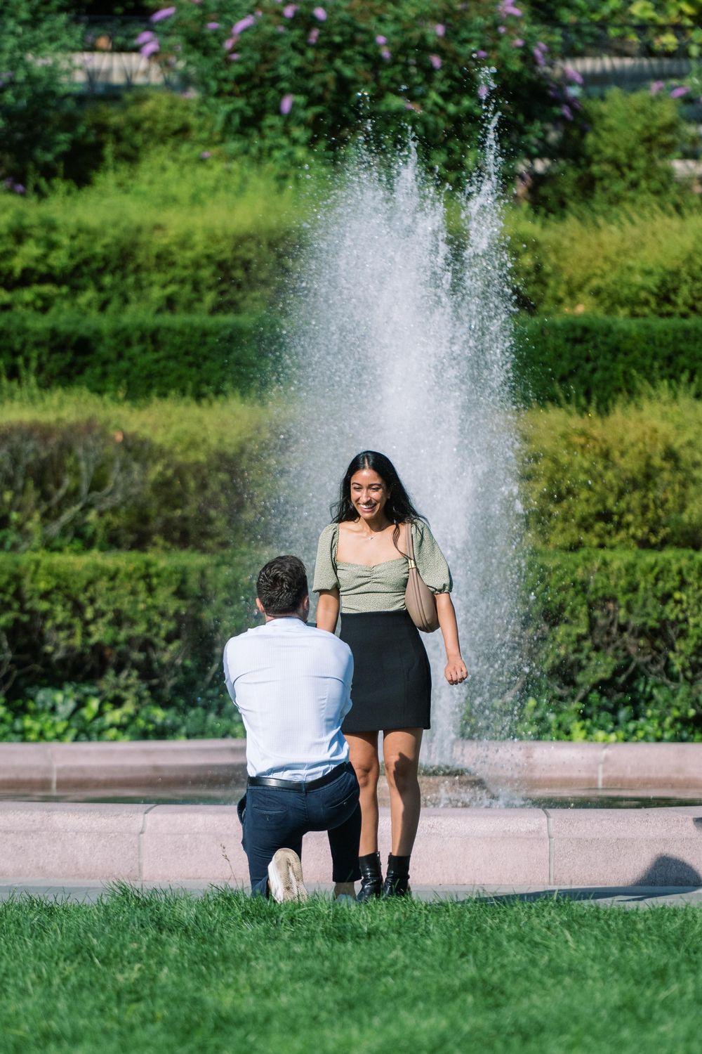 Two people pose together by a water fountain in a peaceful garden setting surprise proposal in conservatory gardens central park.