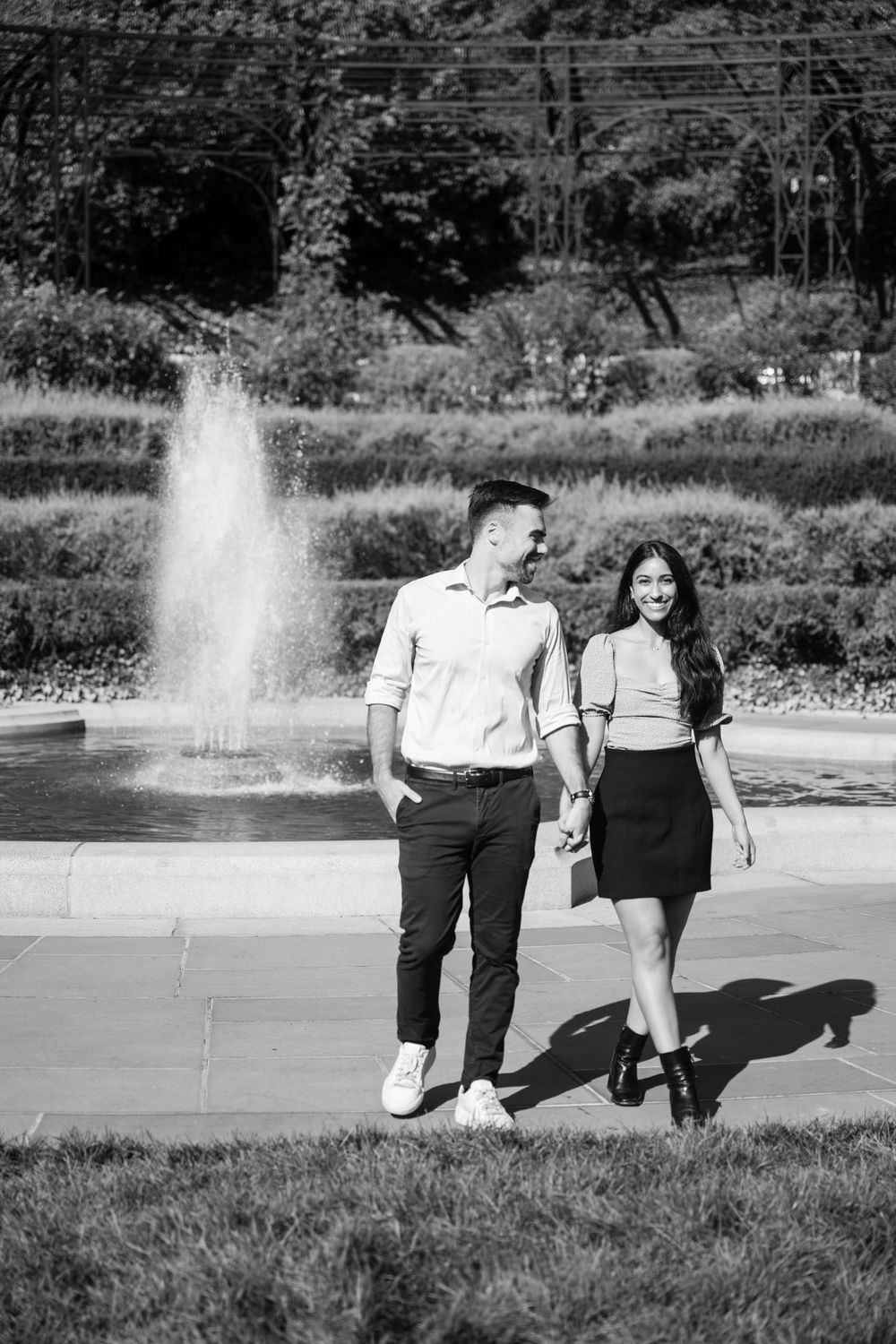 A black and white photo of two people by a fountain in an urban park setting.