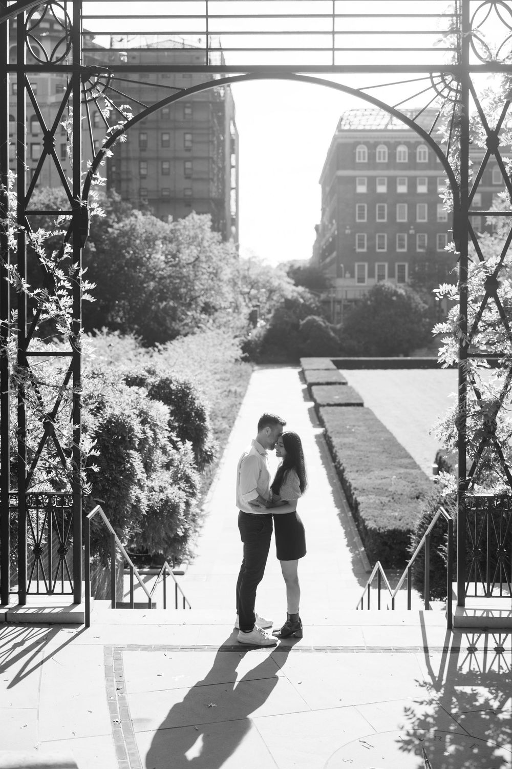 Black and white photo series showing a couple embracing on a walkway with city buildings in the background in conservatory gardens central p