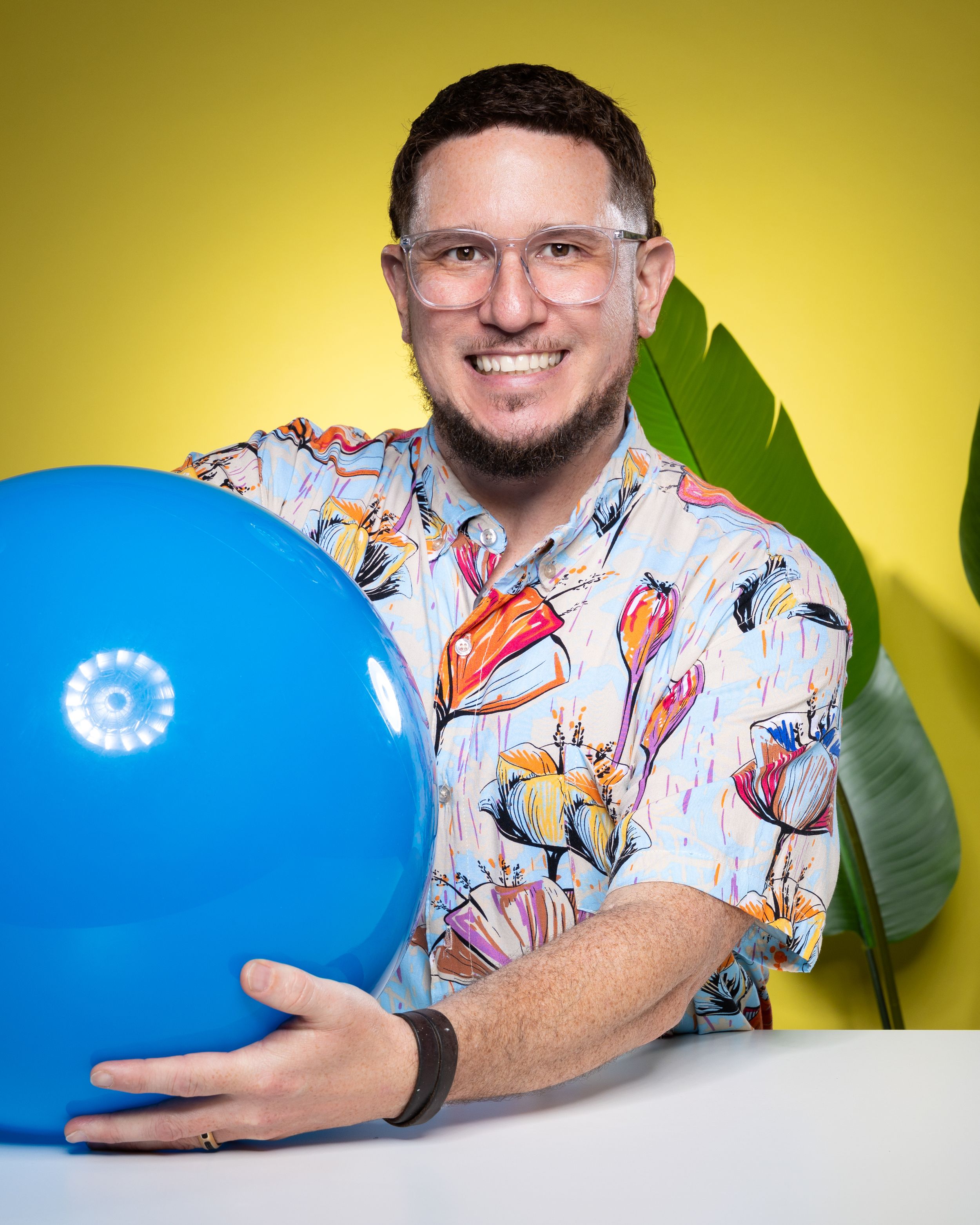 Person in floral print shirt holding blue balloon against yellow background with tropical leaf accent.