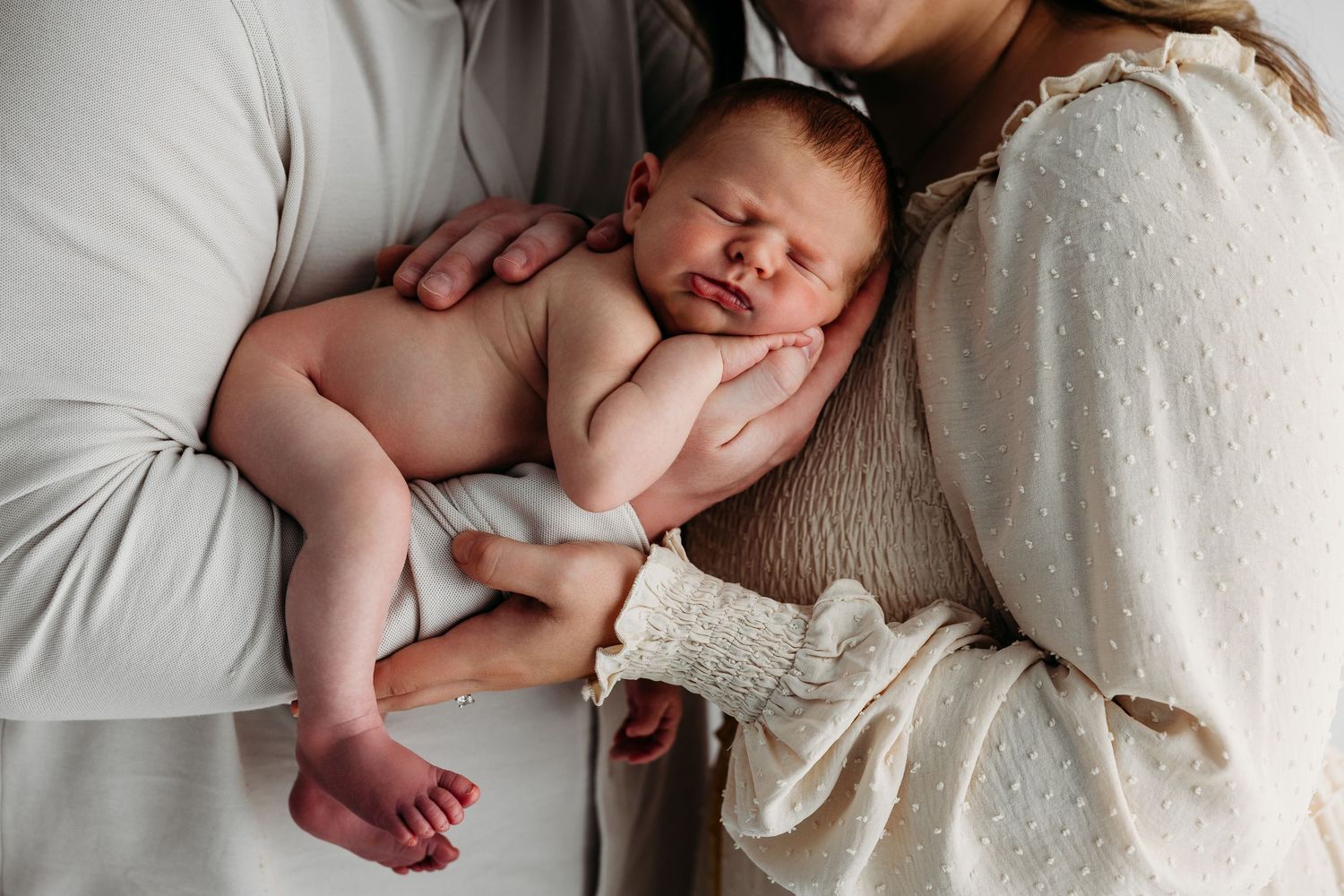 Newborn baby lying on soft white blanket while being gently held during professional photography session.