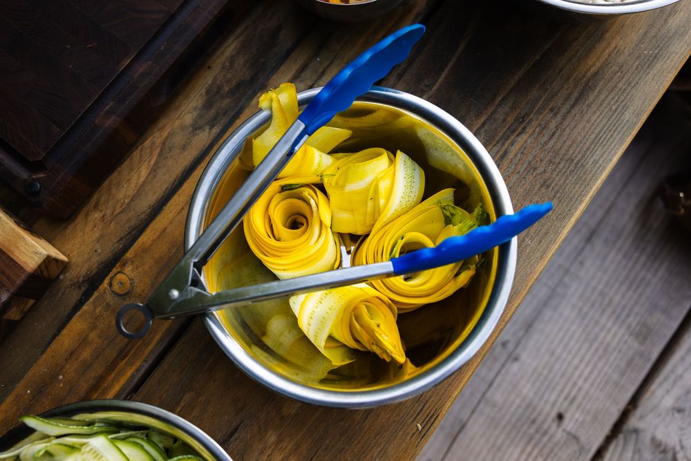Yellow napkins and blue utensils in a metal serving bowl on wooden surface.