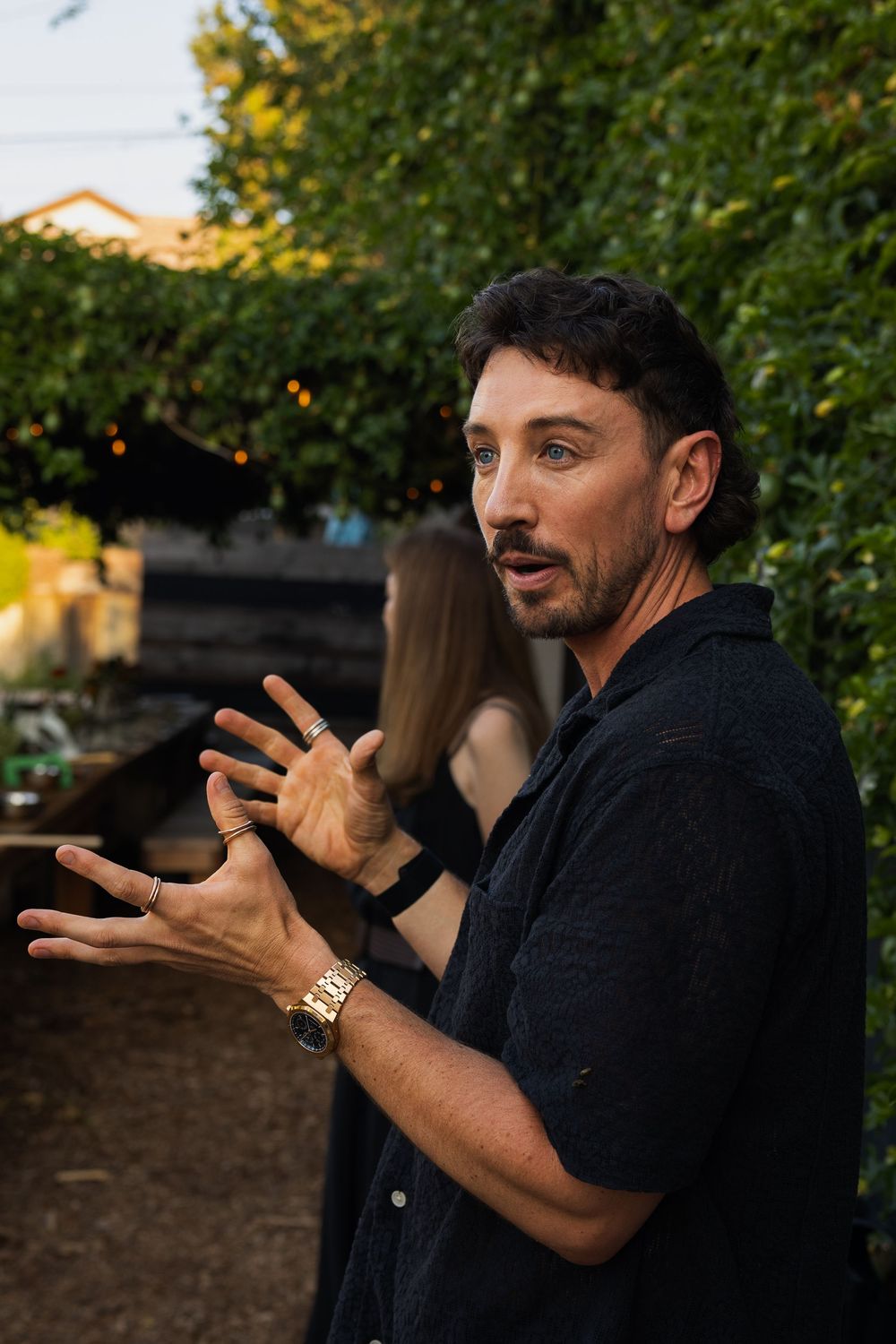 Person gesturing while speaking outdoors with greenery in background.