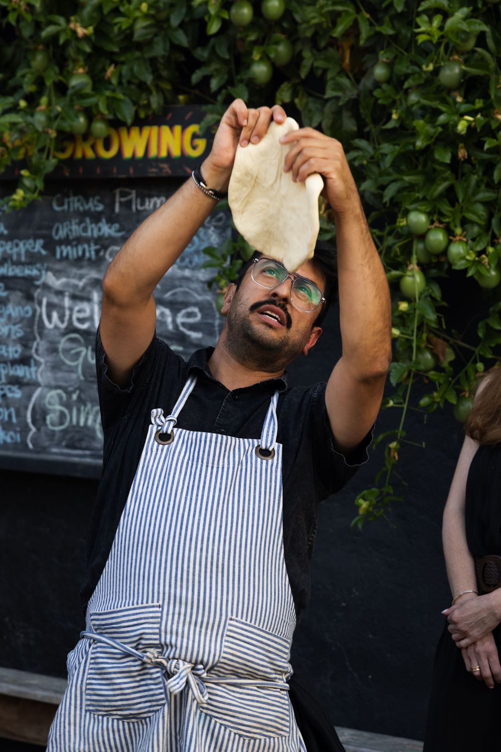 Chef in striped apron stretches pizza dough against dark background with chalkboard menu.