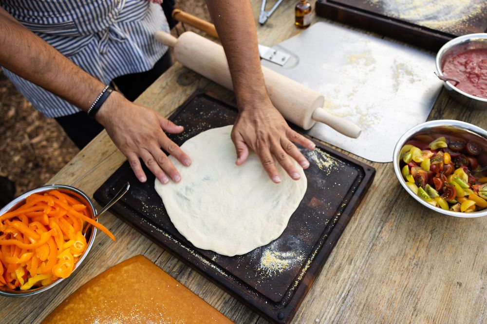 Hands preparing pizza dough on wooden surface with colorful ingredients in bowls.