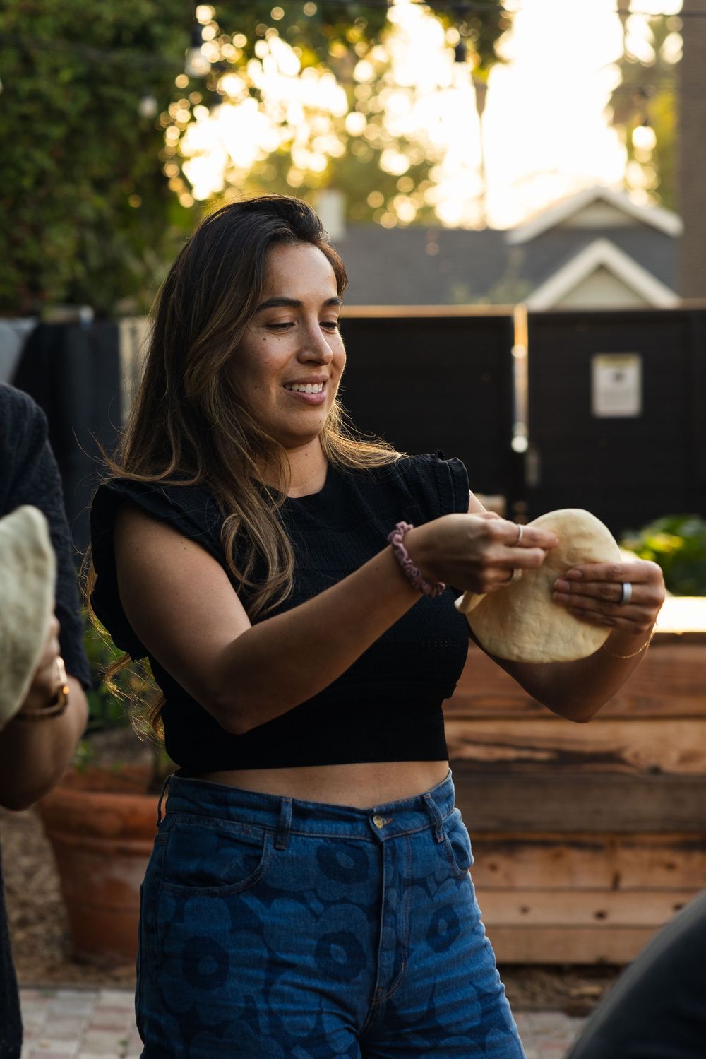 Person stretches pizza dough outdoors during golden hour with wooden tables in background.