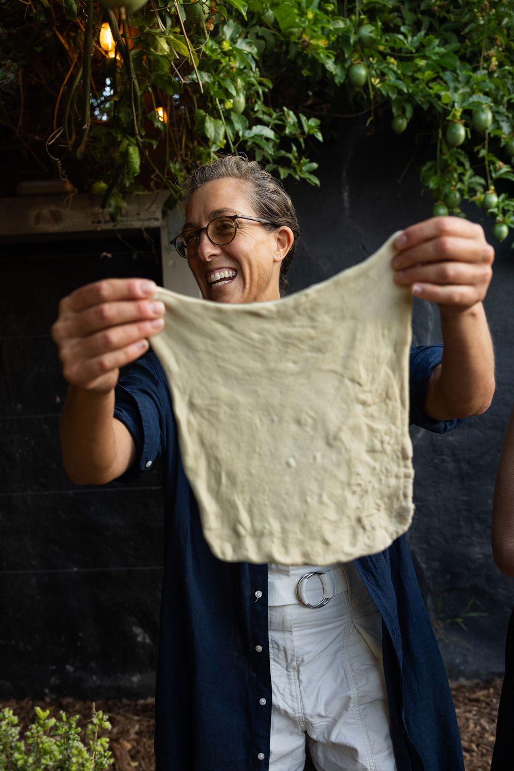 Person stretches pizza dough against dark background with hanging plants and string lights.