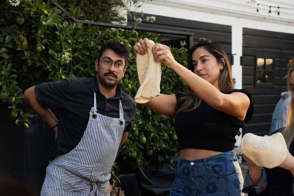 Multiple people stretching pizza dough outdoors during evening light.