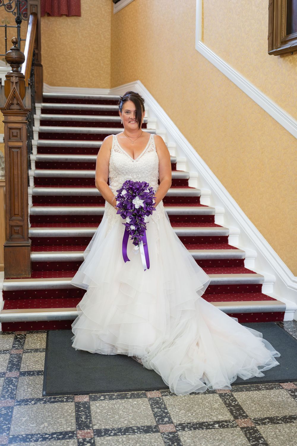 A flowing white wedding dress with purple bouquet photographed on red carpeted stairs.