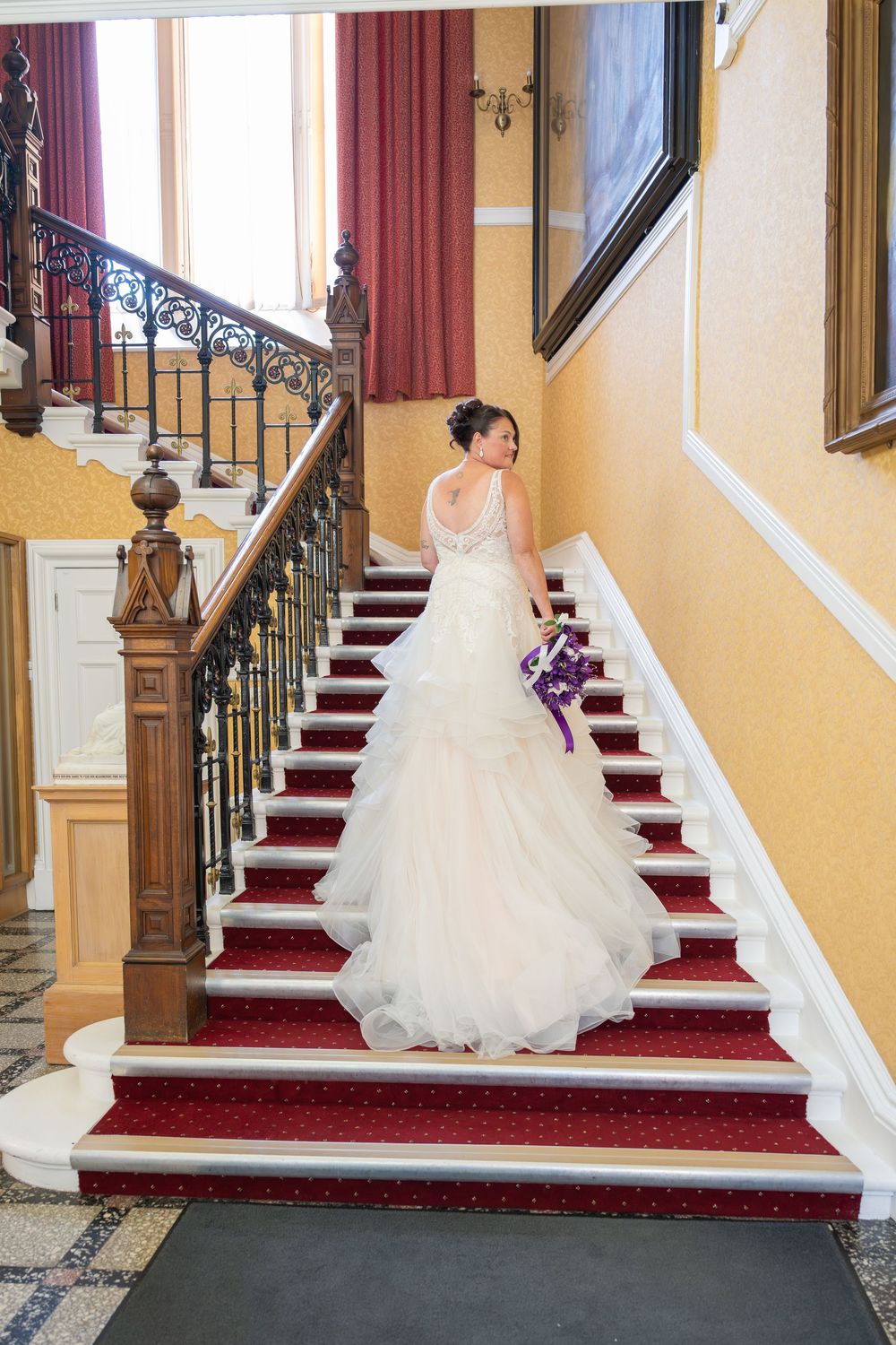 A bride in a white ballgown ascends red carpeted stairs in an elegant venue.