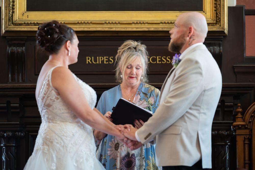 Wedding ceremony participants gather in a formal room with gold-framed artwork.