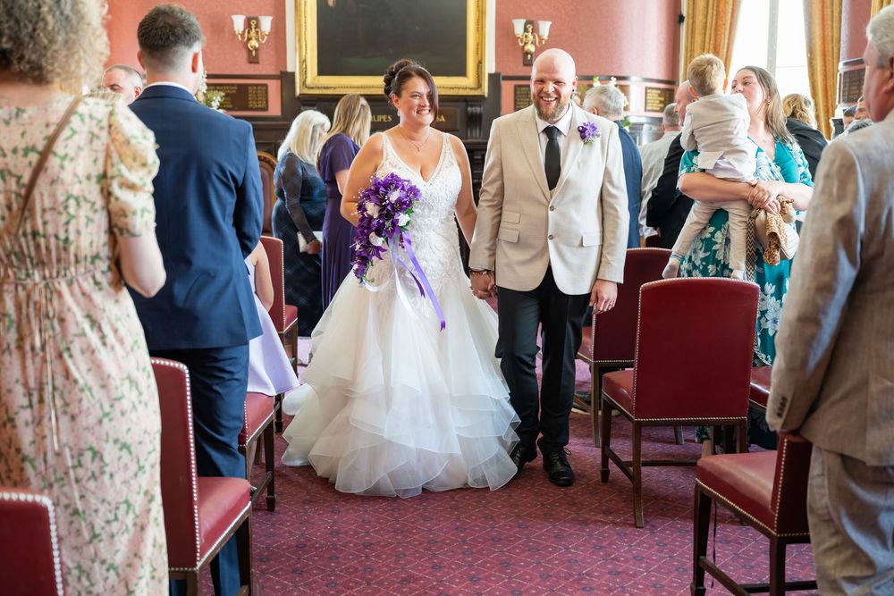 Wedding party walking down aisle in historic venue with red carpet and wooden chairs.