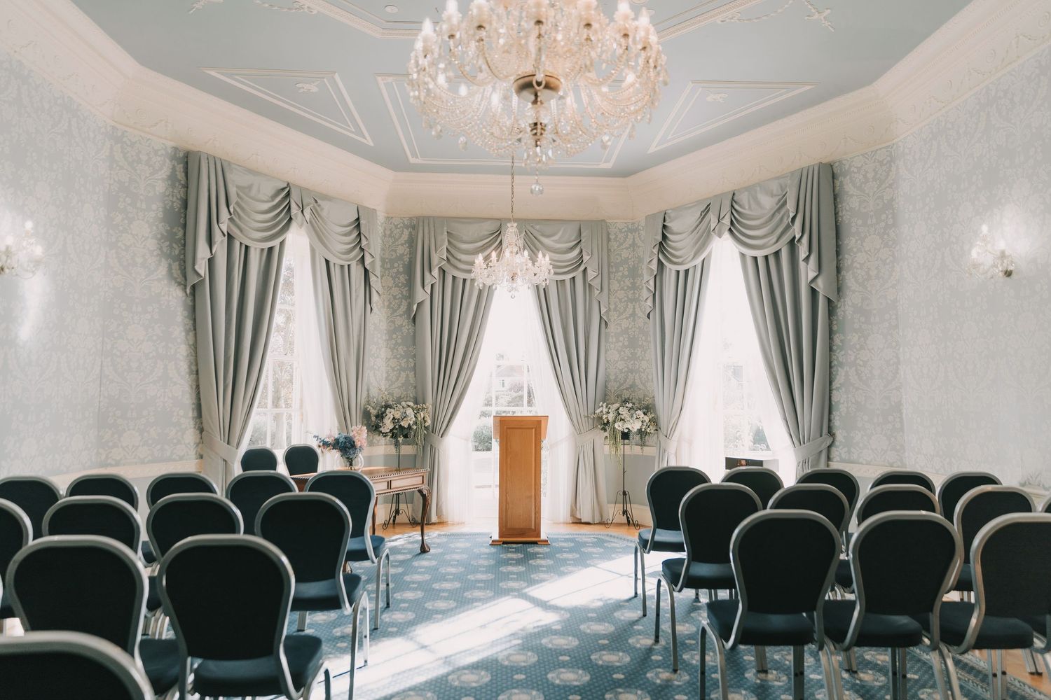 Elegant wedding venue with crystal chandelier, white walls, and rows of black chairs arranged for a ceremony.