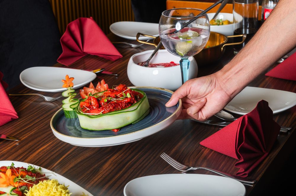 A hand places a plate of beautifully garnished food on a dark wooden restaurant table with red napkins.