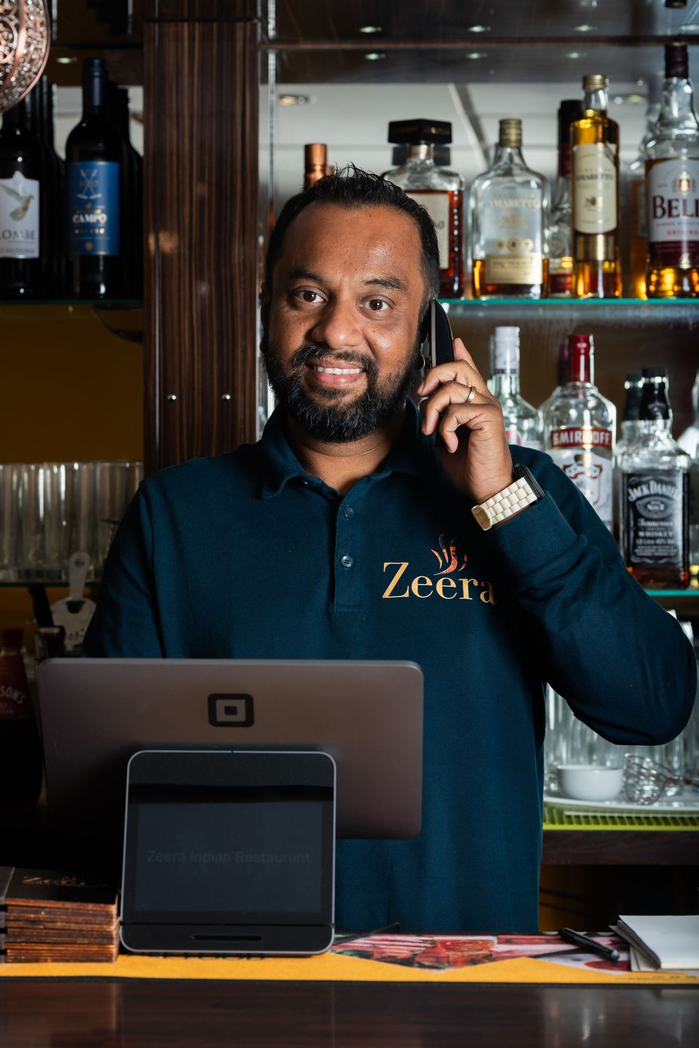A person working at a bar counter with a laptop and phone, surrounded by liquor bottles in the background.