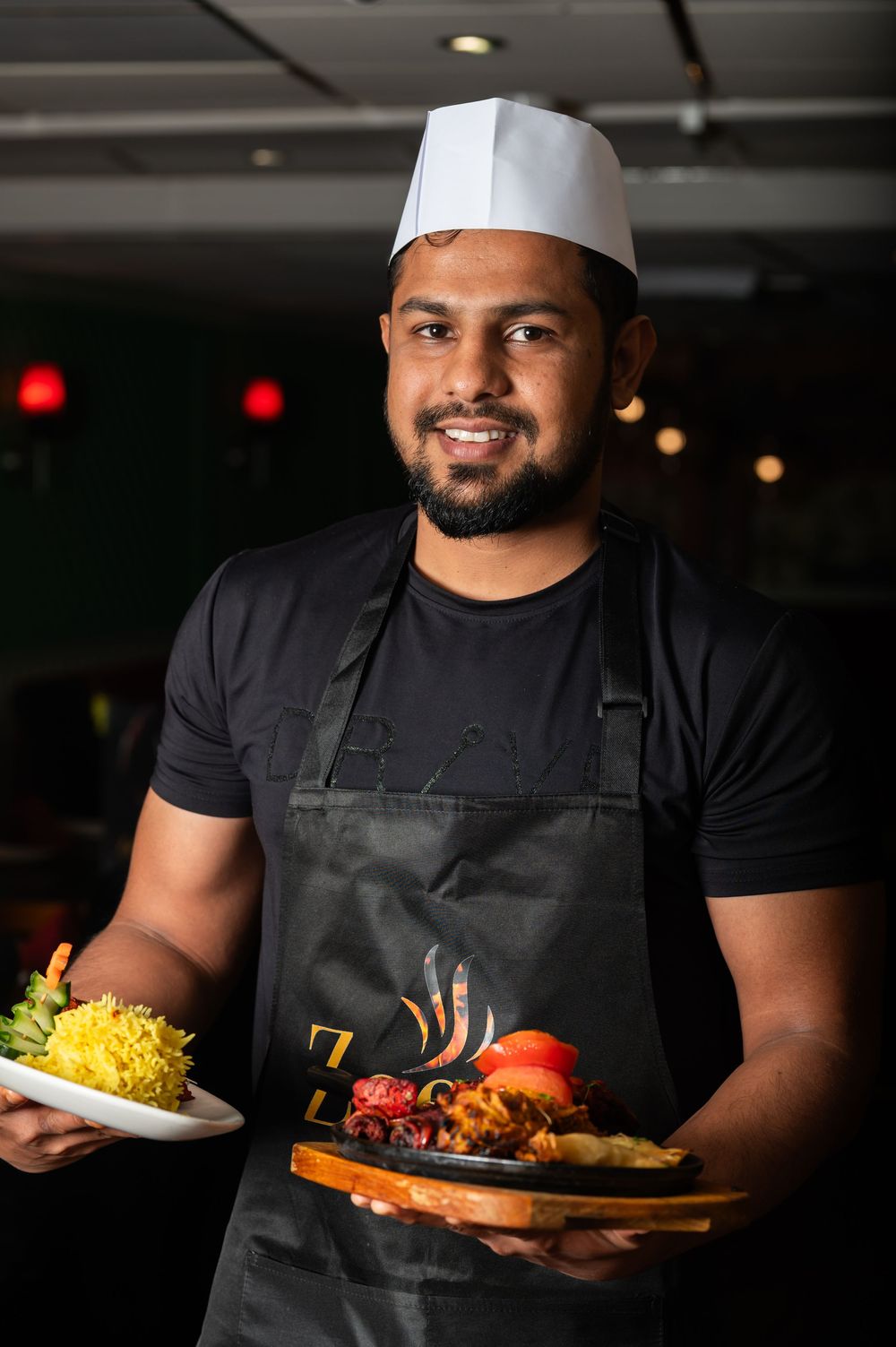 Chef in black apron and hat serves colorful plated dishes in dimly lit restaurant setting.