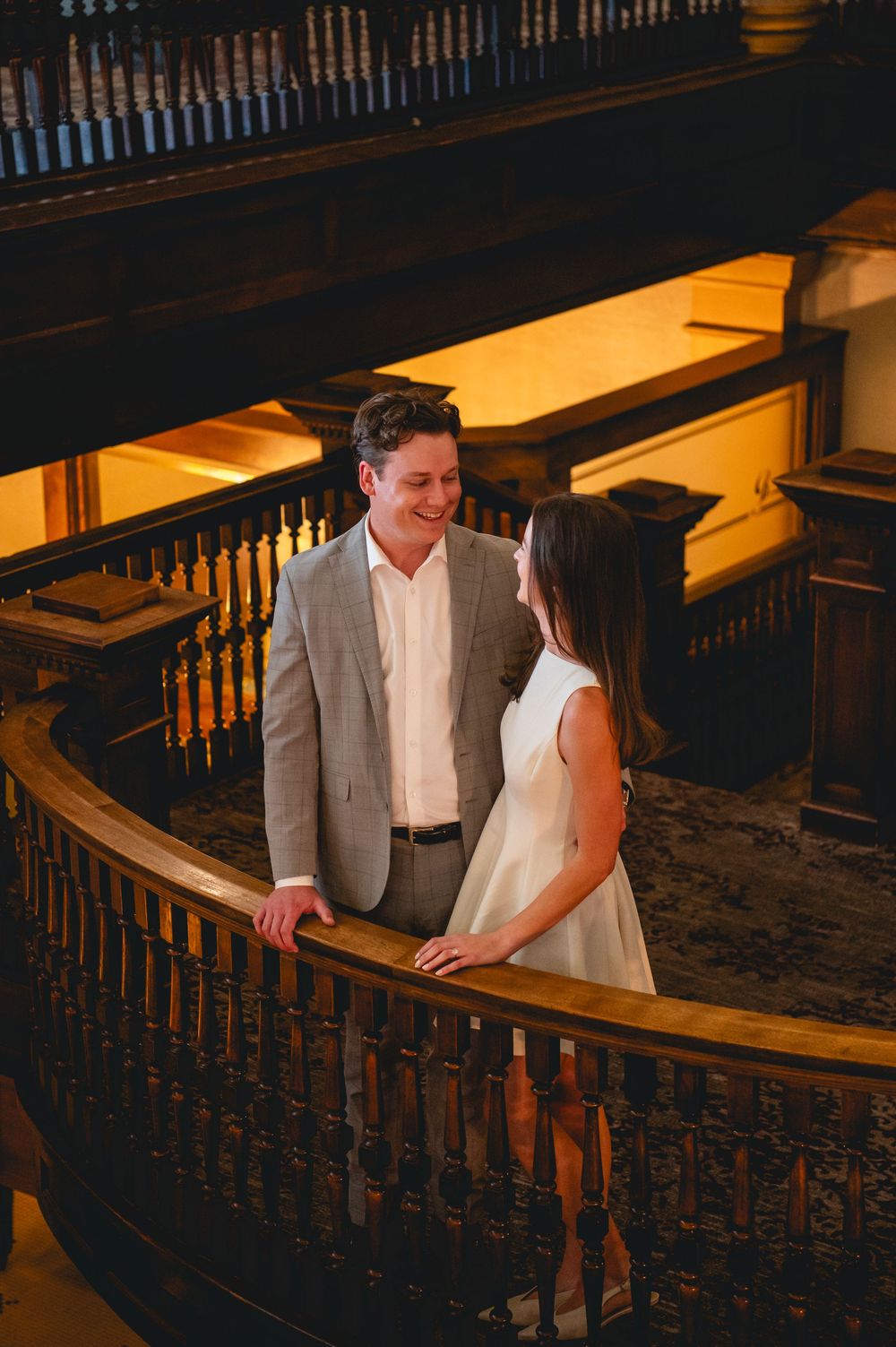 Couple shares a romantic moment on an elegant wooden staircase with ornate railings in warm lighting.