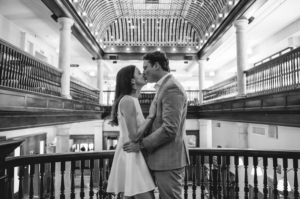 A romantic couple embraces in a modern shopping mall with white railings and escalators in the background.