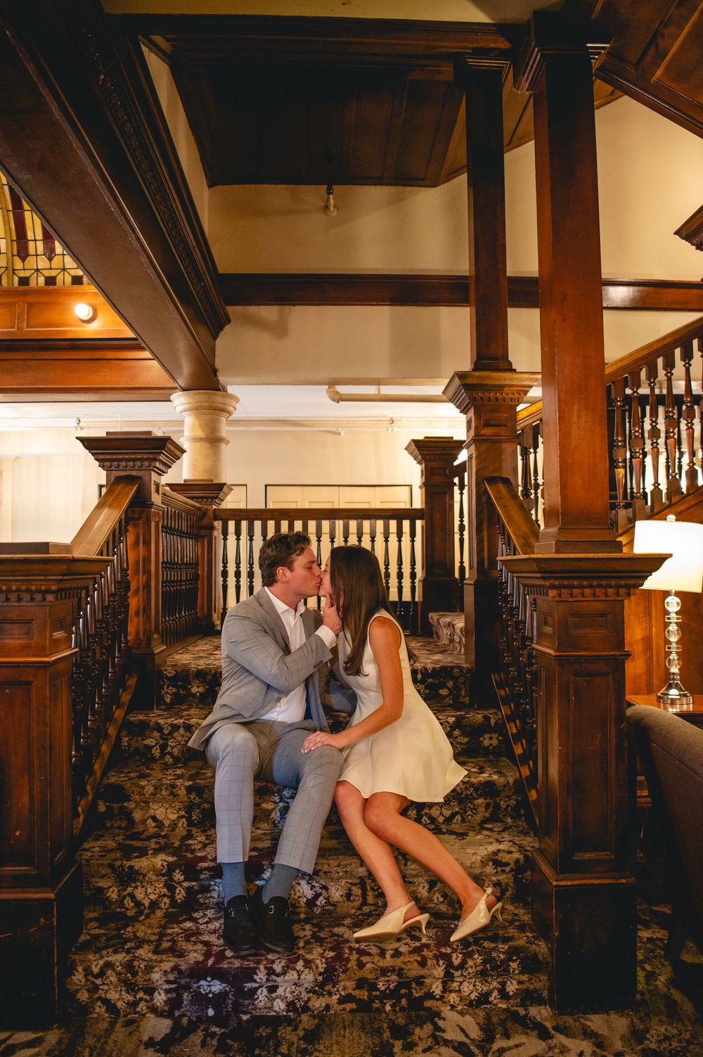 Couple sits together on carpeted stairs surrounded by vintage wooden columns and banisters in historic building.