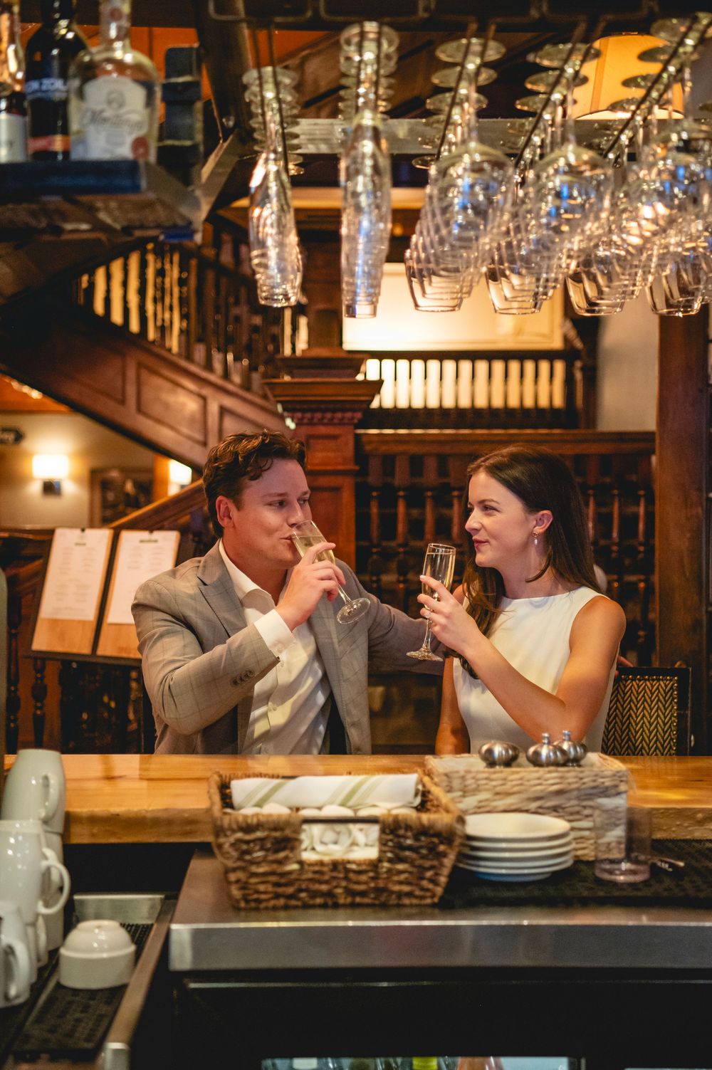 Couple enjoying wine and conversation at a cozy restaurant bar with hanging glassware and warm lighting.